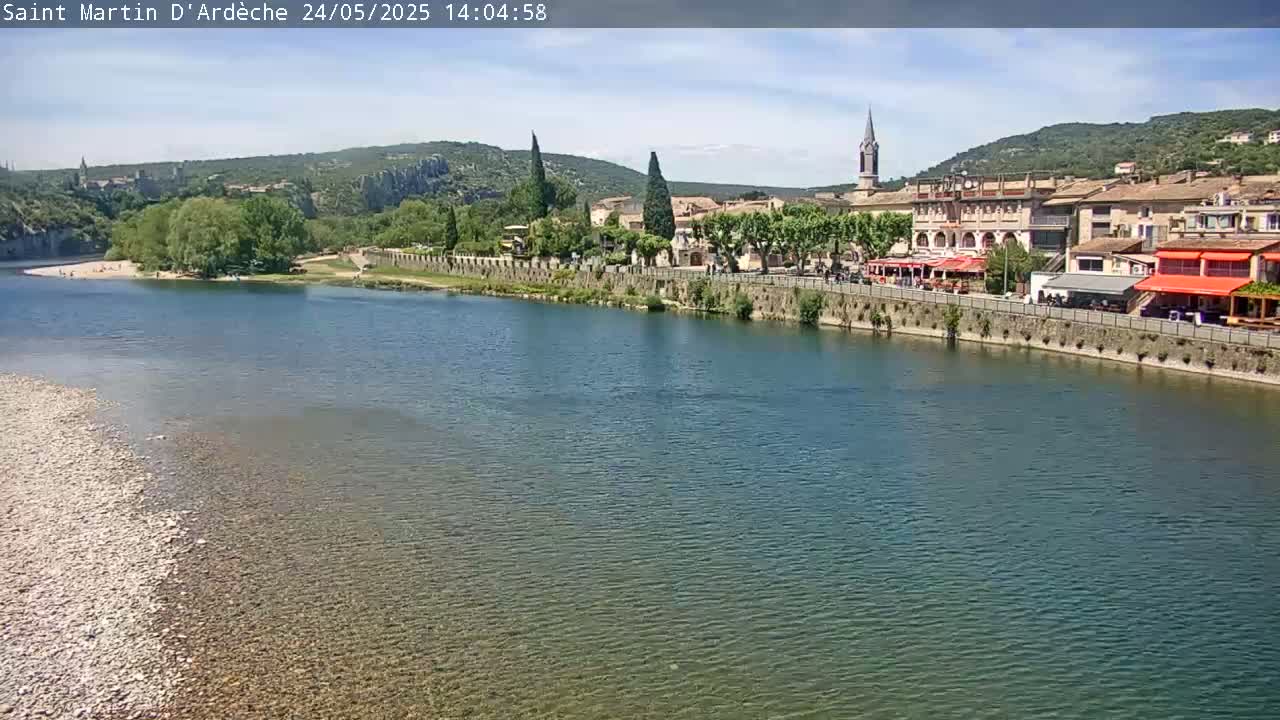 A river flows past a village nestled beside hills under a mostly sunny sky.