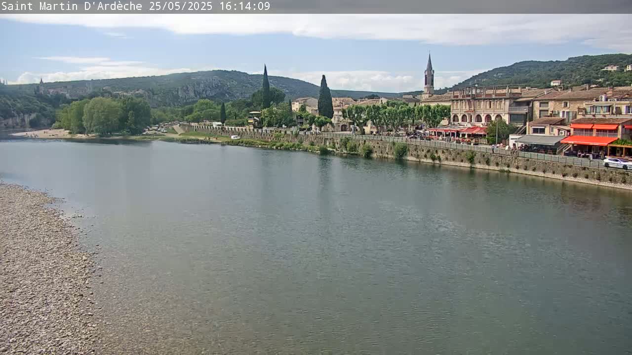 A river flows past a village nestled amongst hills under a partly cloudy sky.