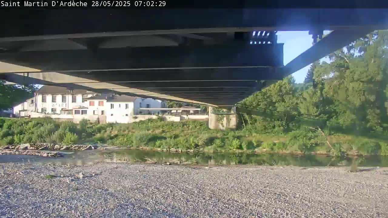 Under a bridge on a sunny day, a calm river flows past a gravel bank and a line of buildings nestled among green trees.