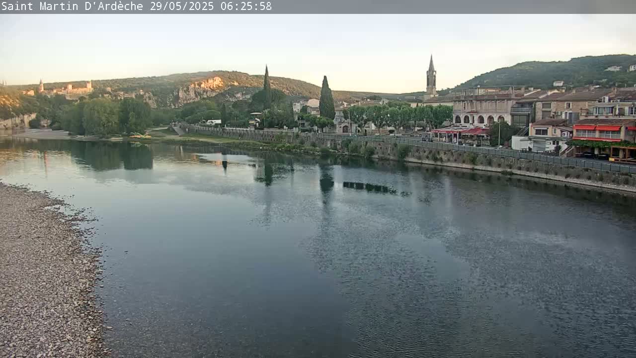 A calm river reflects a town nestled amongst hills under a clear sky.