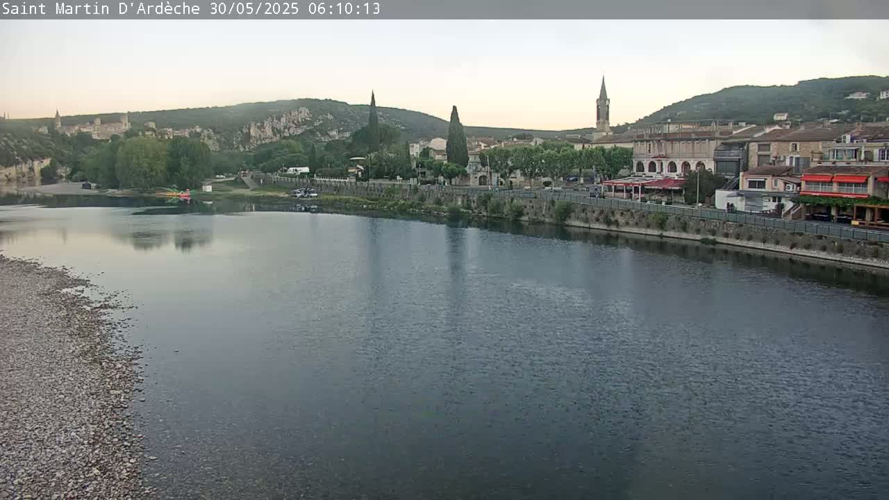 A calm river reflects a small town nestled against hills under a pale, overcast sky.