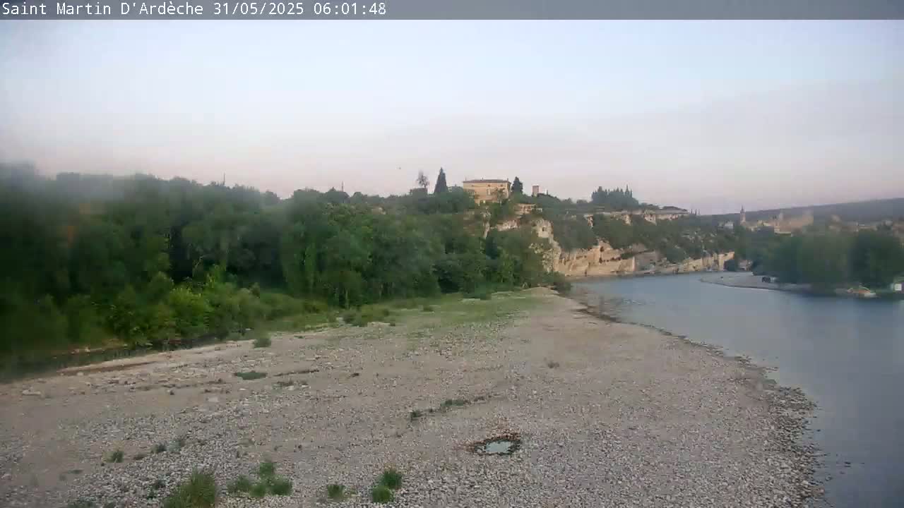 A wide, rocky riverbank stretches towards a calm river, beyond which sits a village nestled against a tree-covered hillside under a hazy sky.