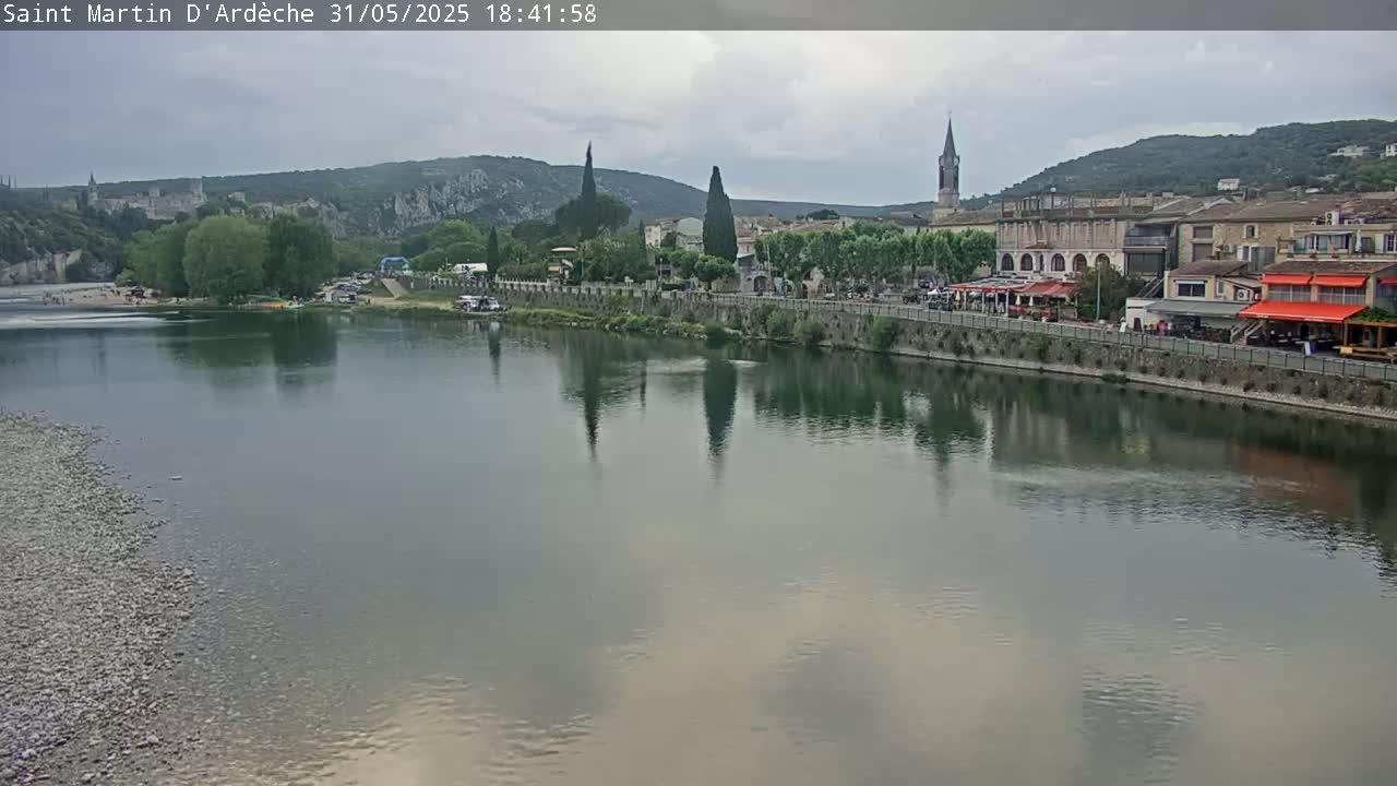 A calm river reflects a town nestled against hills under a cloudy sky.