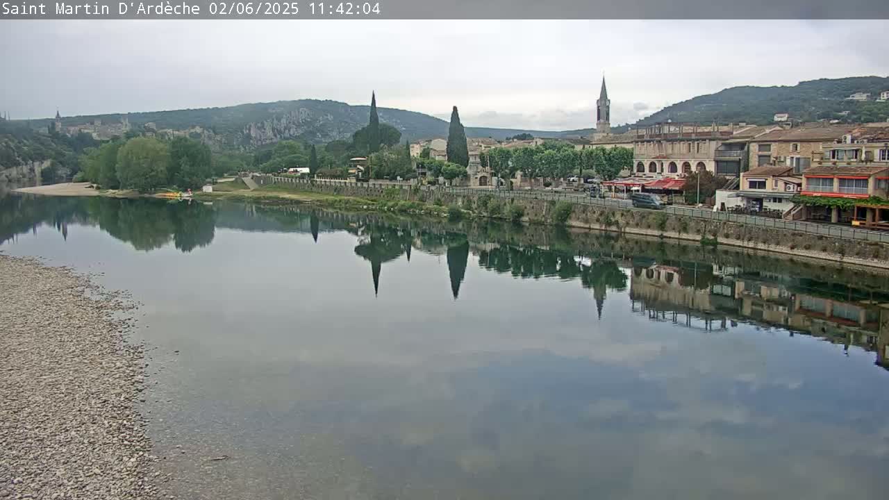 A calm river reflects a town nestled against hills under an overcast sky.