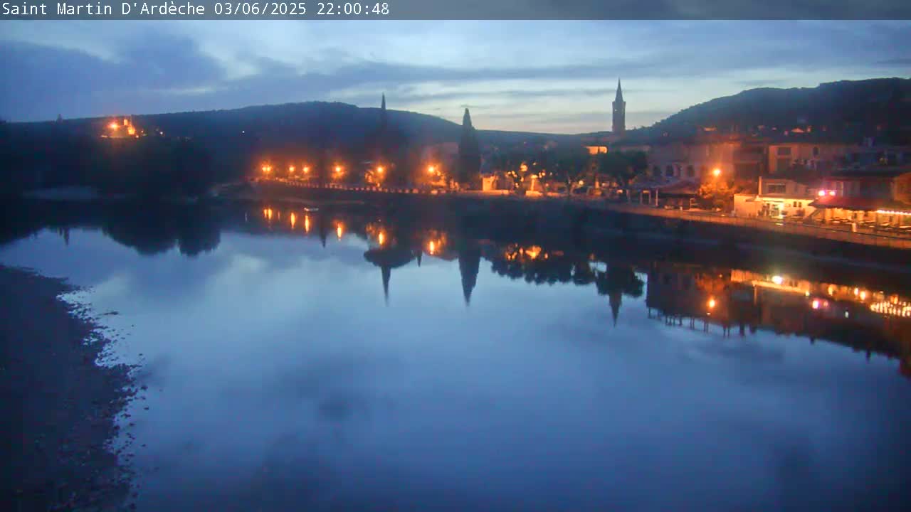A nighttime view of a town with lights reflecting on a calm river under a twilight sky.