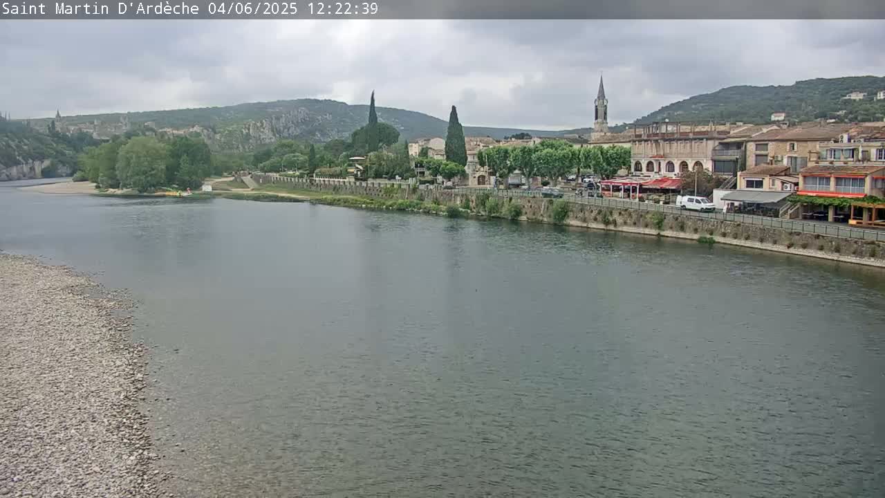 A river flows past a town nestled amongst hills under a cloudy sky.