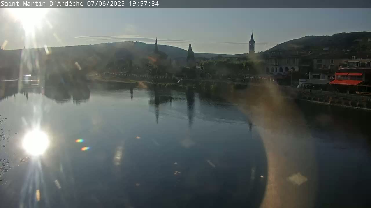 A calm river reflects the setting sun and the silhouette of a town with several church steeples on the opposite bank.