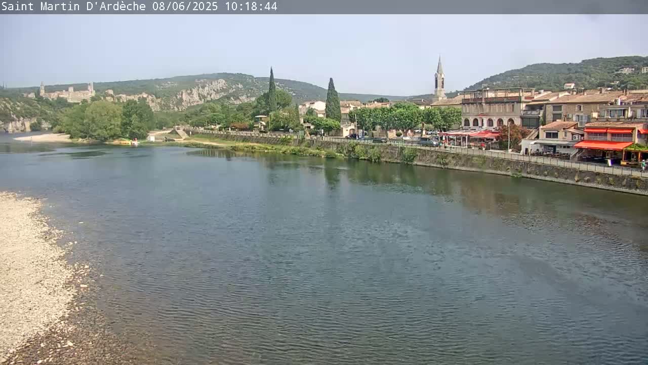 A calm river flows past a village nestled against hills under a clear sky.