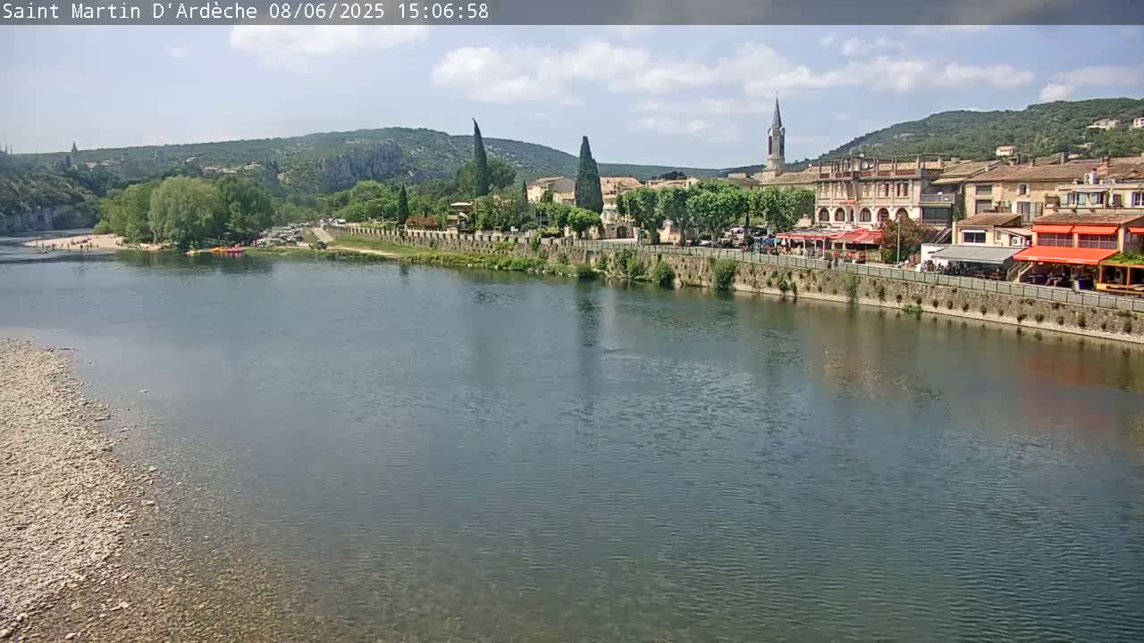 A calm river flows past a town nestled in a valley under a partly cloudy sky.