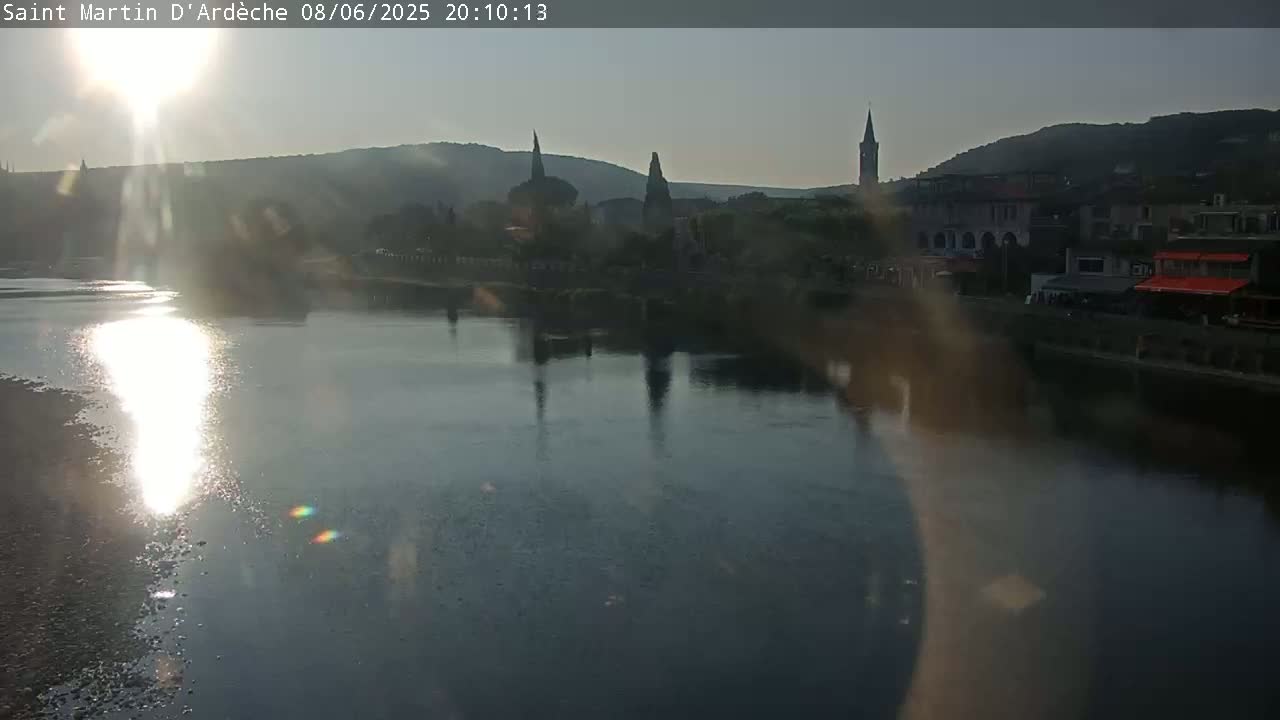 A calm river reflects the setting sun and the silhouettes of buildings and church steeples on the opposite bank.