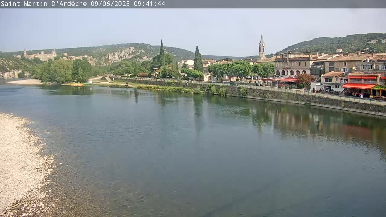 A calm river reflects a village nestled amongst hills under a clear sky.