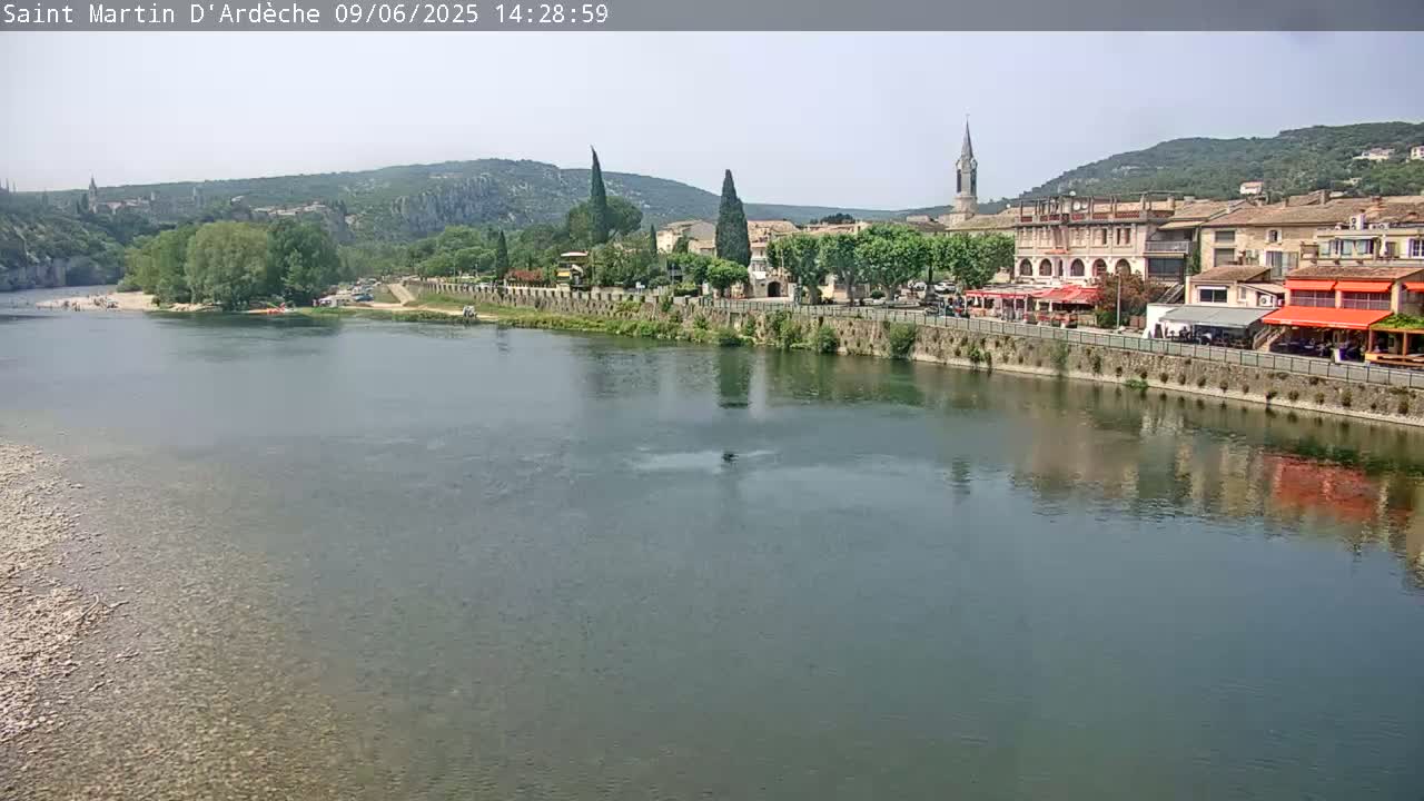 A calm river reflects a village of buildings and trees under a sunny sky, nestled between hills.