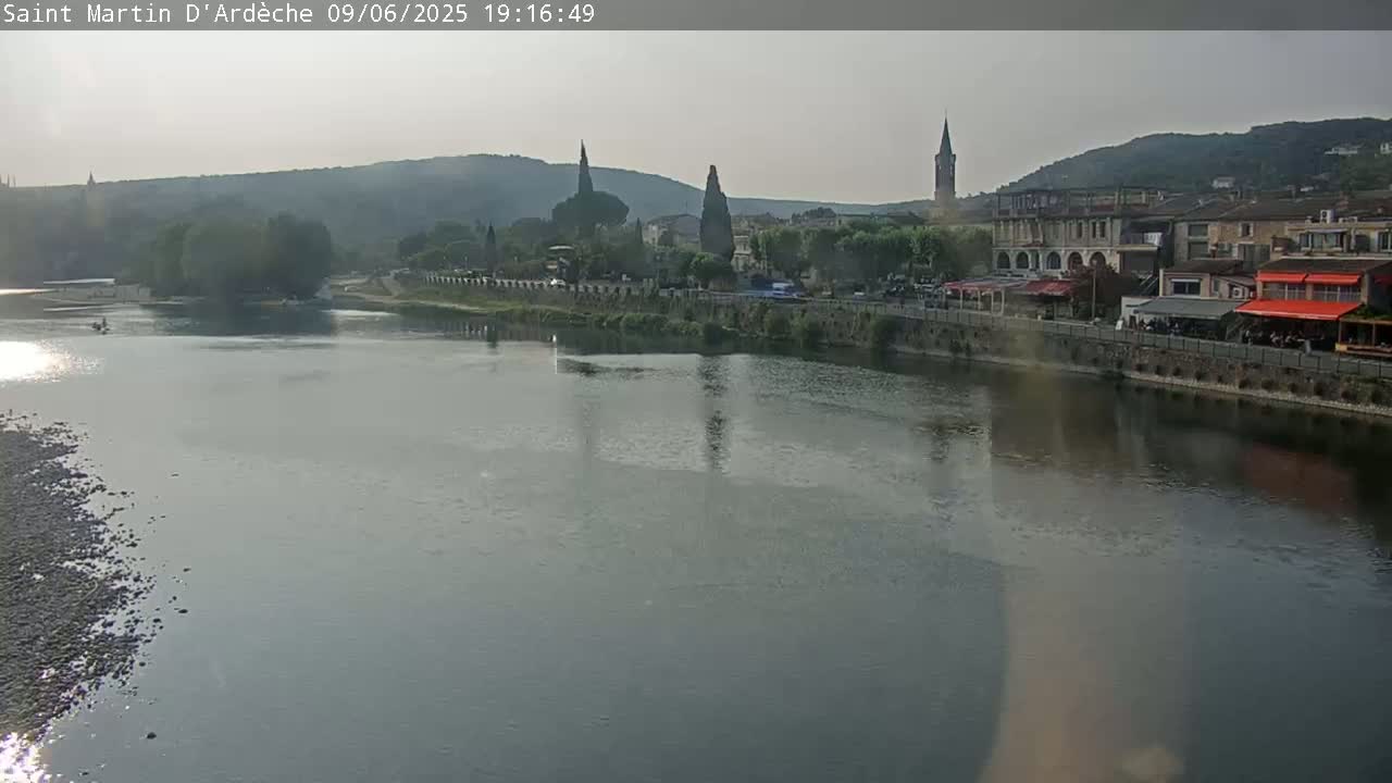 A calm river reflects a town with several buildings, including church steeples, under a hazy sky.