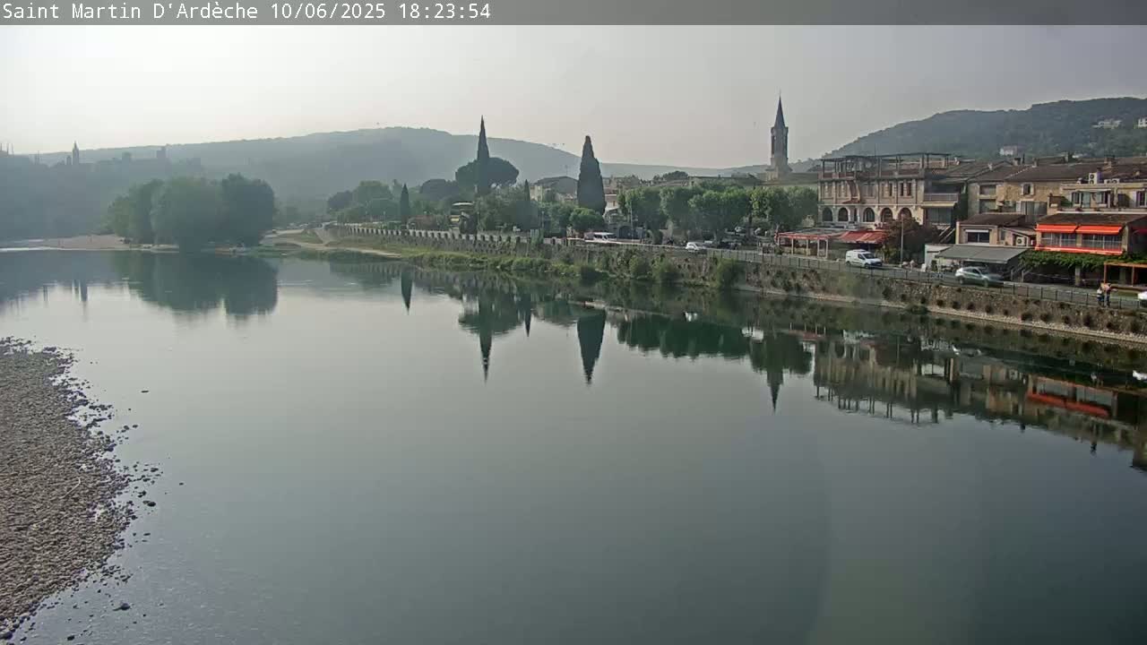 A calm river reflects a town with several buildings and church spires, under a hazy sky.