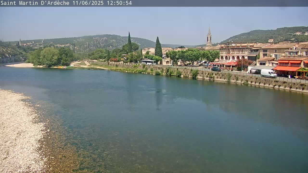 A river flows past a village nestled in a valley under a sunny sky.