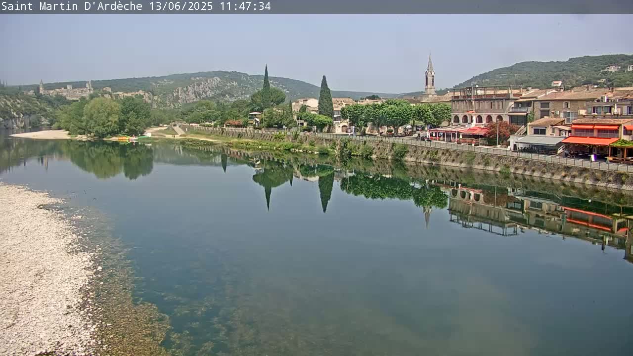 A calm river reflects a village nestled among hills on a sunny day.