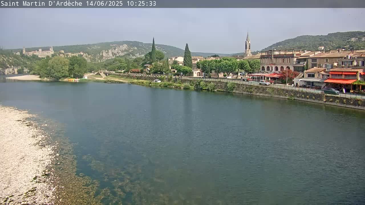 A river flows past a village nestled beside hills under a clear sky.