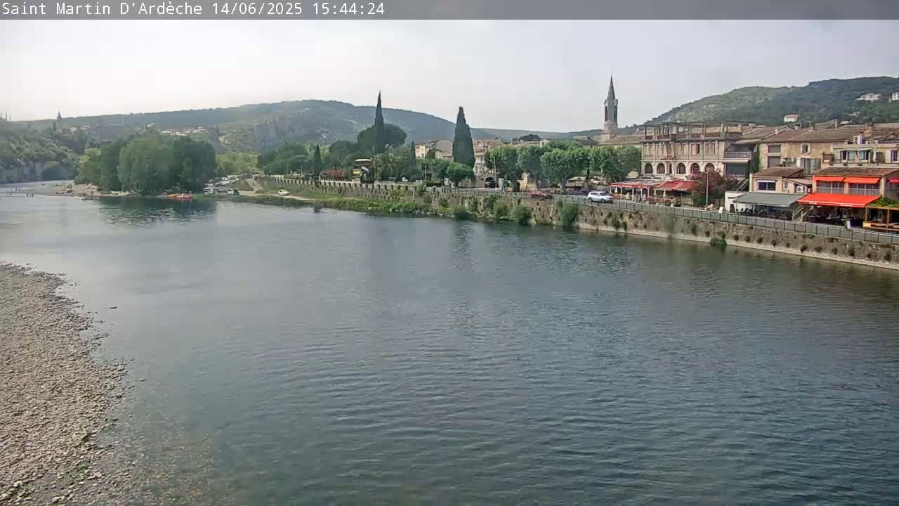 A river flows past a town nestled in a valley under a partly cloudy sky.