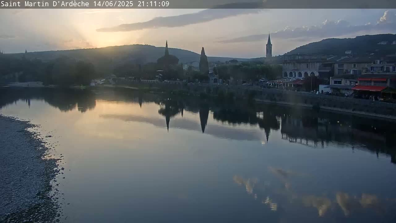 A river flows past a town nestled in a valley under a partly cloudy sky.