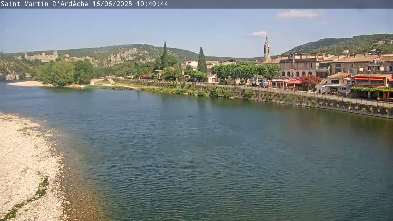 A river flows past a village nestled beside hills under a clear, sunny sky.