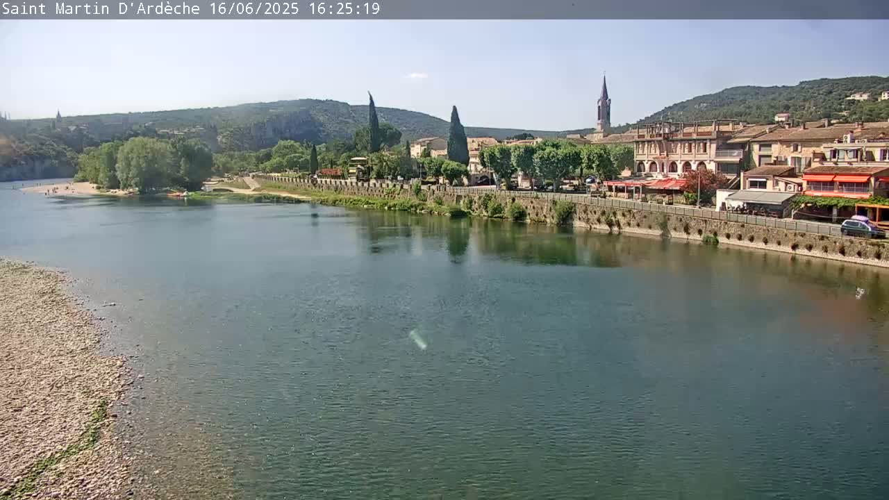 A river flows calmly past a village nestled beside hills under a clear, sunny sky.