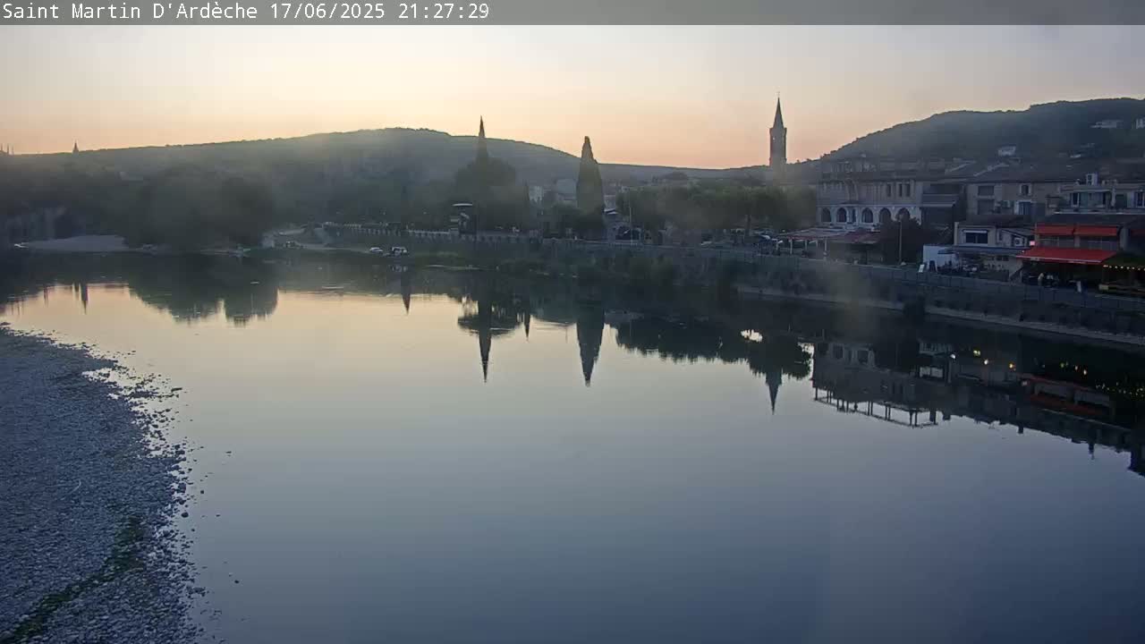 A calm river reflects a town's buildings and church steeples at twilight under a hazy sky.