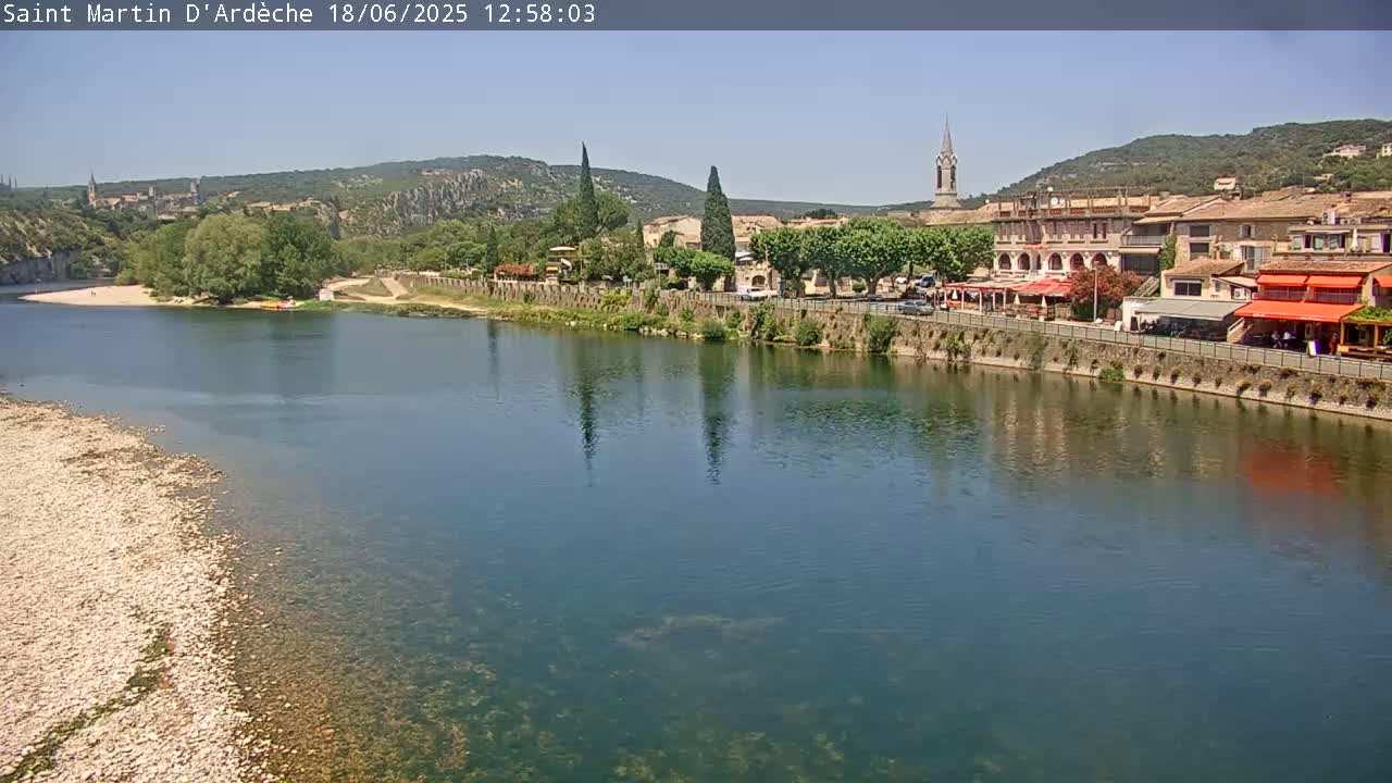 A river with a calm surface reflects a village of buildings and trees under a clear, sunny sky.