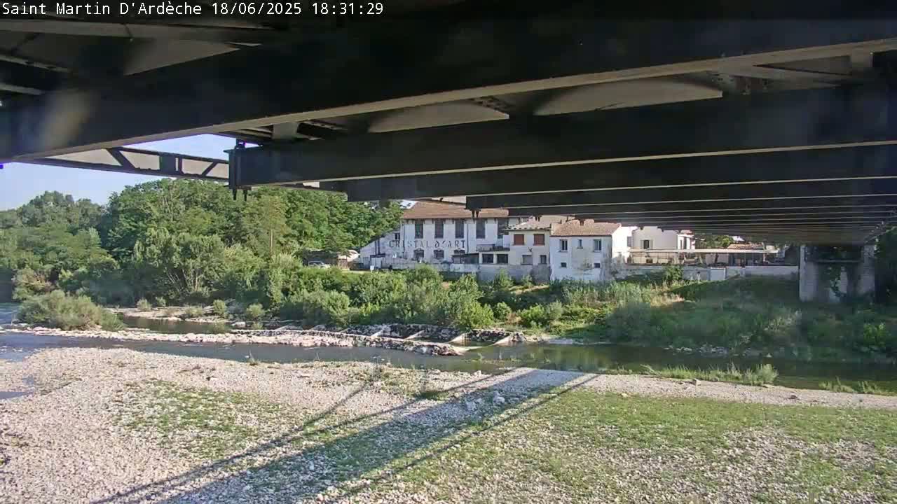 A view from under a bridge on a sunny day shows a river, a gravel riverbank, lush green vegetation, and buildings in the distance.