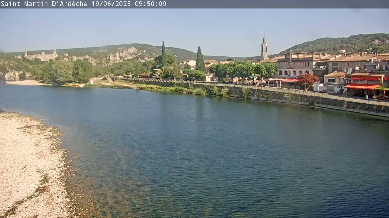 A quaint town sits on the bank of a calm, blue river under a clear, sunny sky, with hills rising in the background.