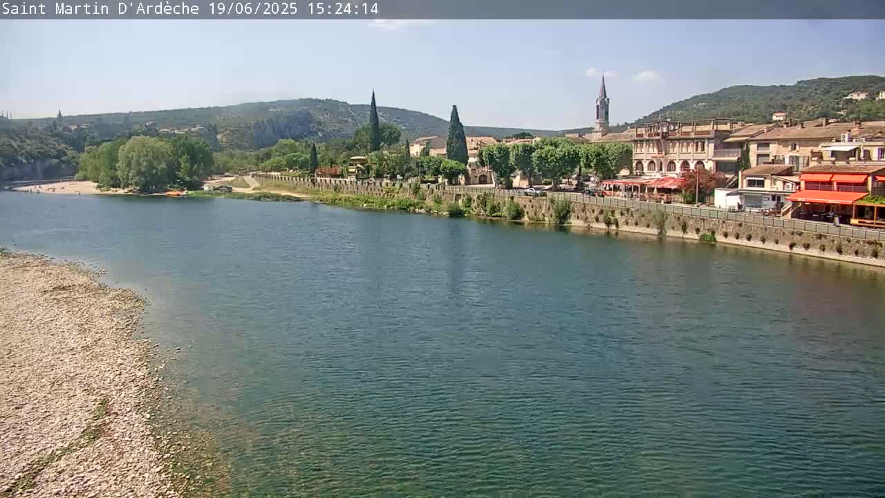 A river flows past a town nestled in a valley under a clear, sunny sky.