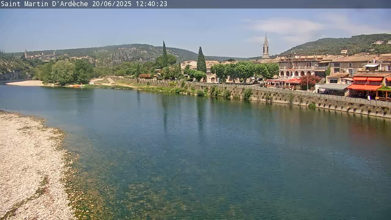 A river flows past a town nestled against a hillside under a clear, sunny sky.