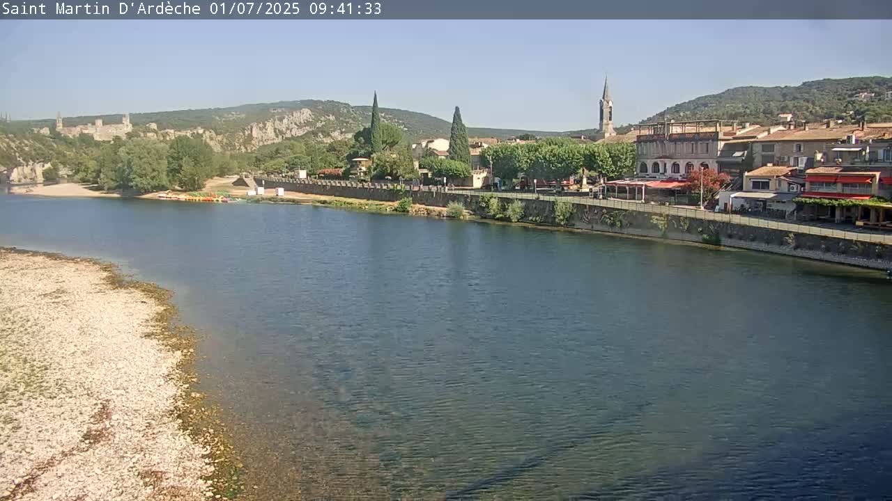 A river flows past a village nestled against a hillside under a clear, sunny sky.