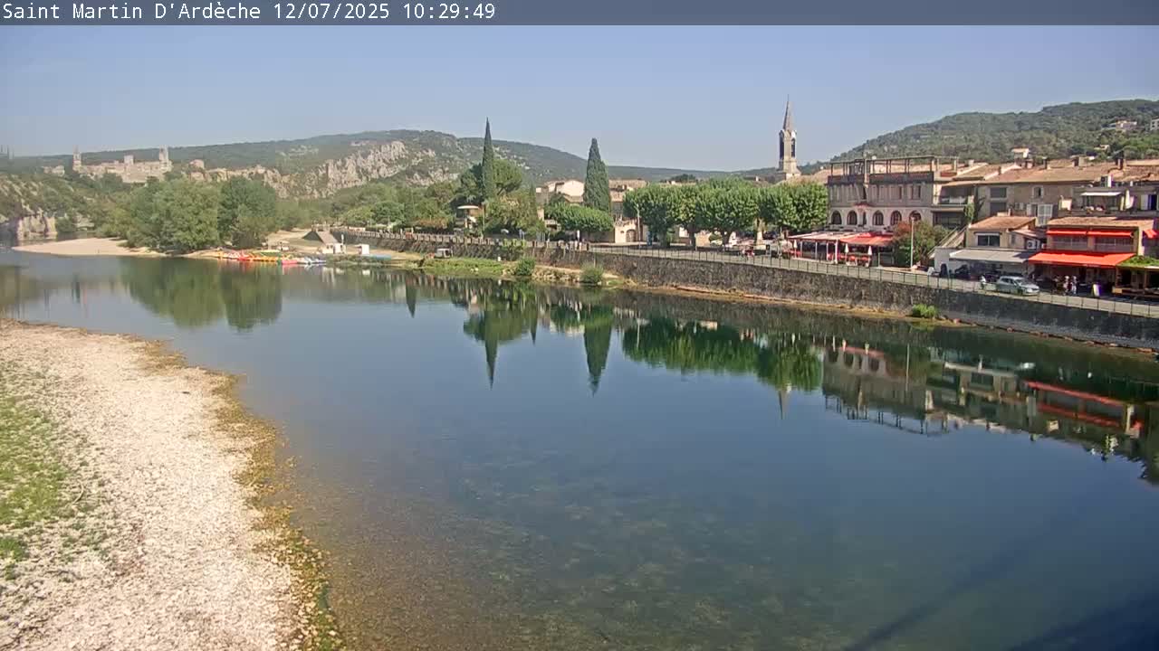 A calm river reflects a village of buildings and trees under a clear, sunny sky.