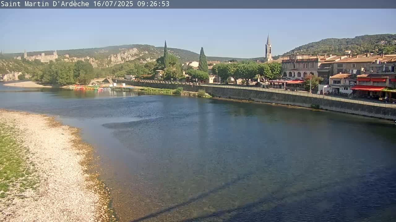 A river flows past a town nestled in a valley under a clear, sunny sky.