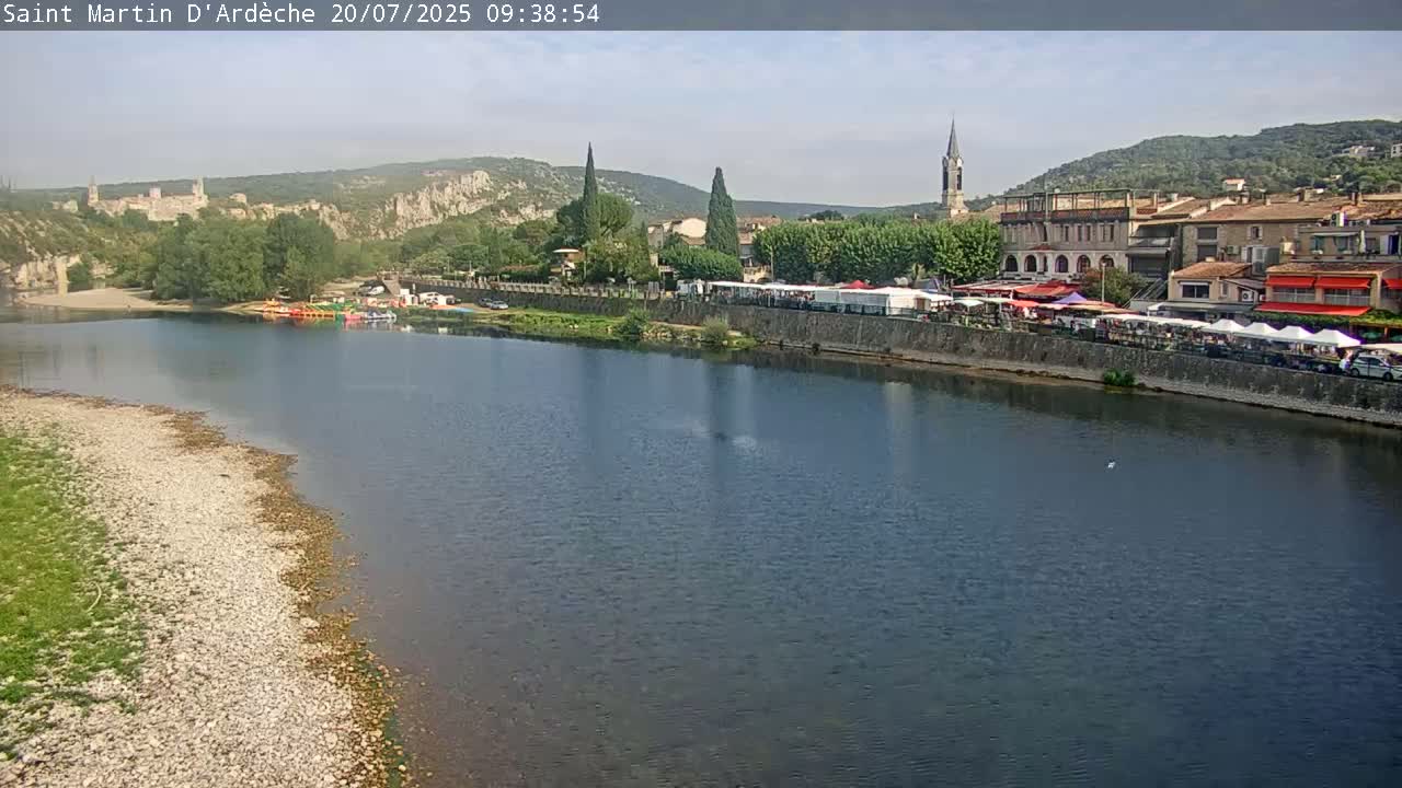 A river flows past a town nestled in a valley under a mostly clear sky, with boats moored along the riverbank and market stalls lining the waterfront.