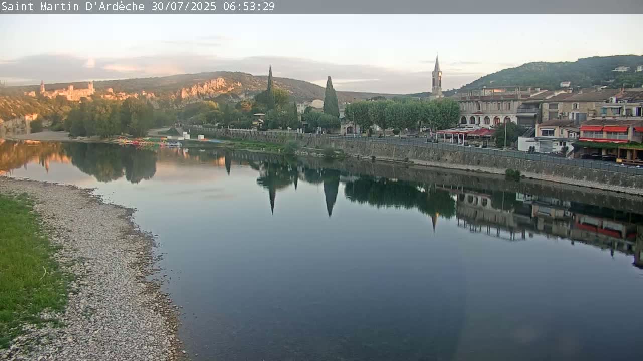 A river flows past a town nestled in a valley under a mostly clear sky, with boats moored along the riverbank and market stalls lining the waterfront.