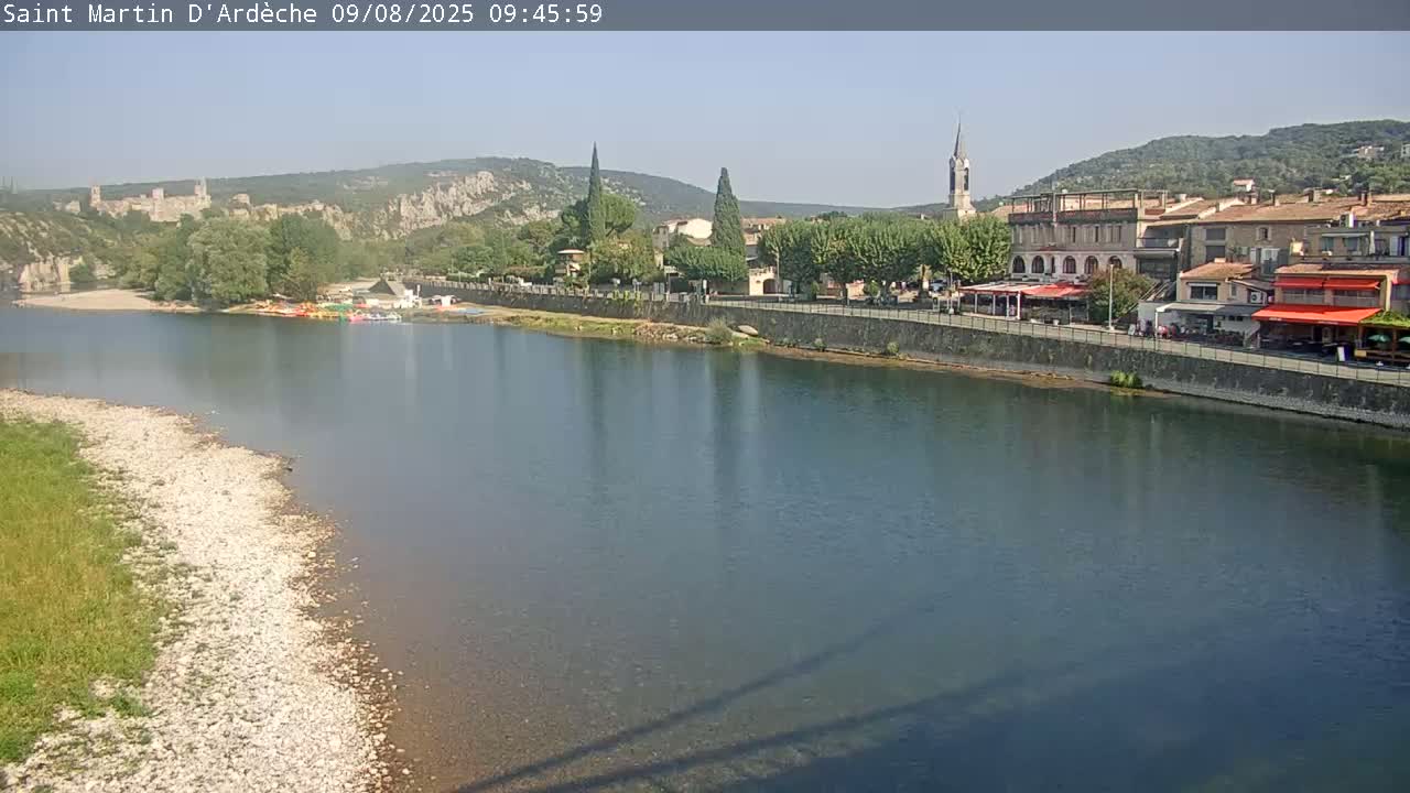 A river flows past a village nestled amongst hills on a sunny day, with boats visible on the riverbank.