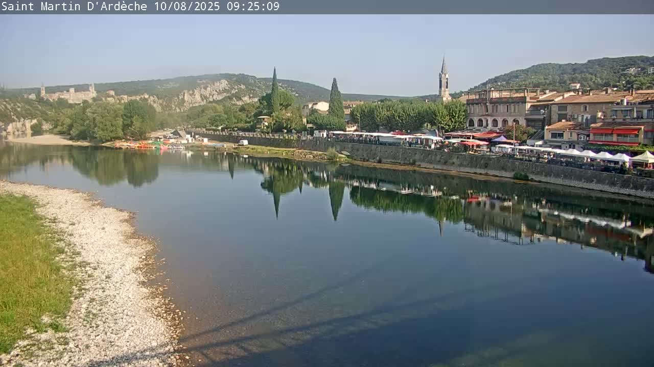 A calm river reflects a town nestled between hills on a clear, sunny day.