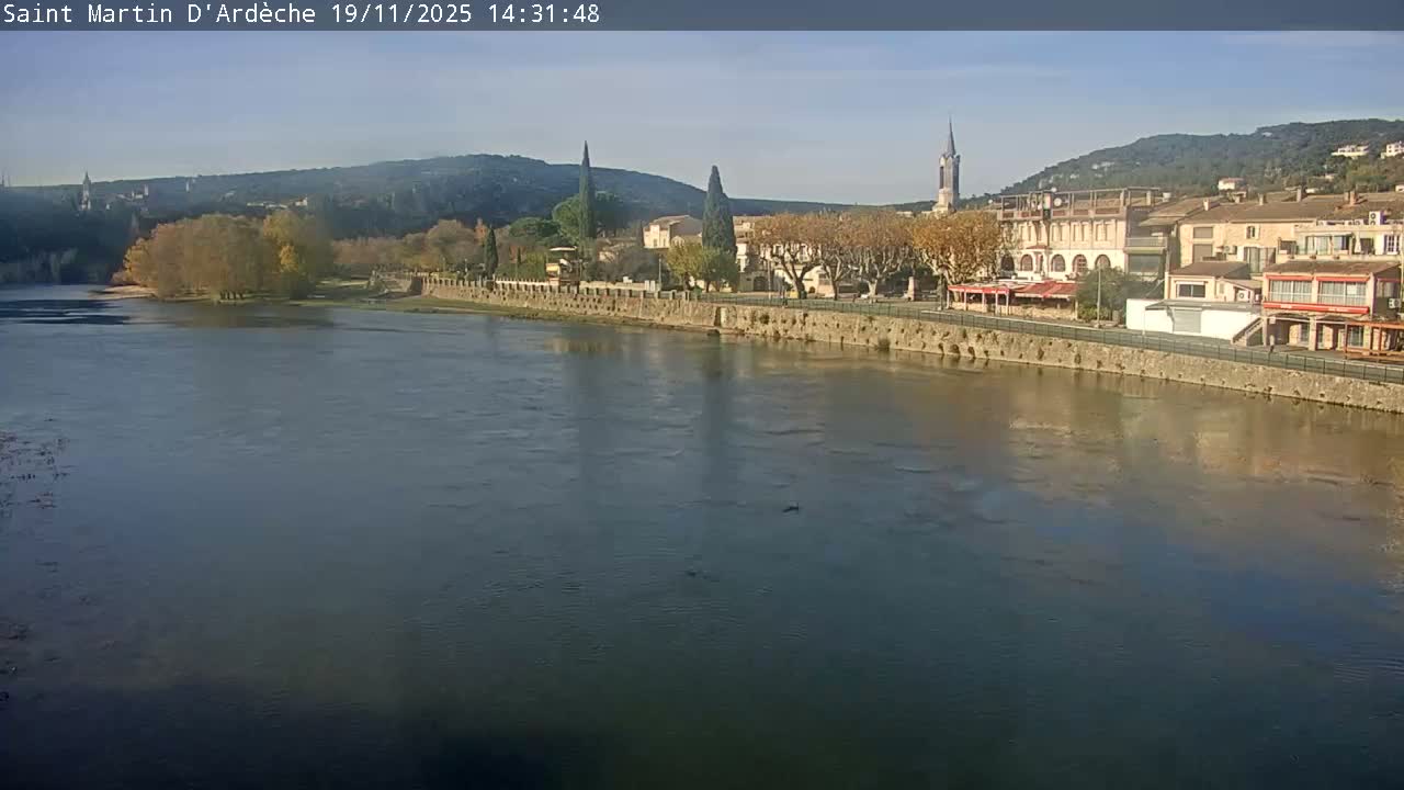 A calm river reflects a town nestled between hills on a clear, sunny day.