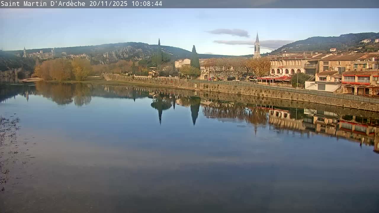 A tranquil river reflects the clear blue sky and a charming town with church spires and autumn-tinged trees nestled against rolling hills, under bright, sunny weather.