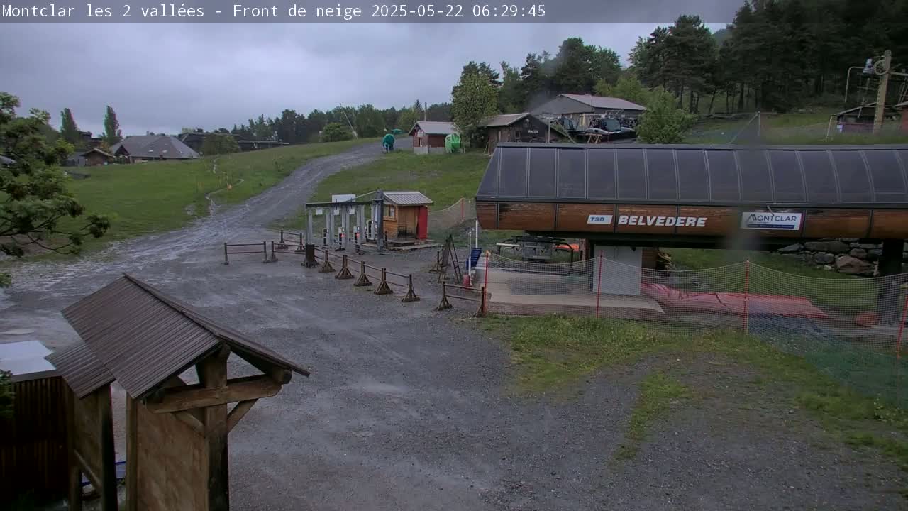 A rainy view of a ski resort base area showing a chairlift station, small buildings, and a gravel path.