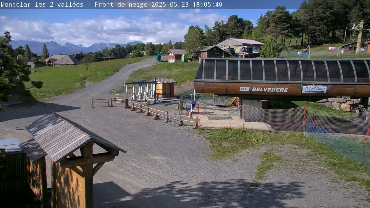 A gravel path leads to a chairlift station in a mountainous area under partly cloudy skies.