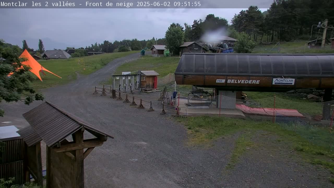 A chairlift station sits on a gravel path amidst green hills and trees under an overcast sky.