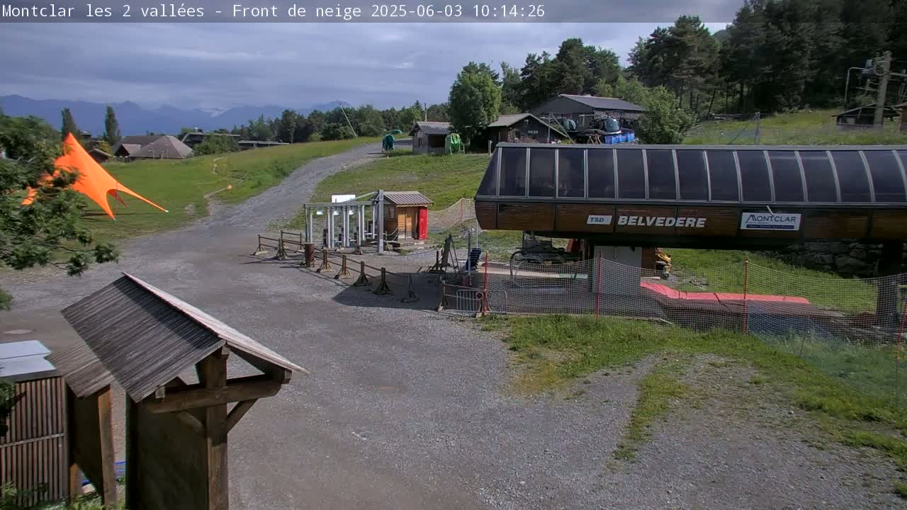 A mostly sunny outdoor scene shows a chairlift base station, a gravel path, some buildings, and an orange canopy, with mountains visible in the background.