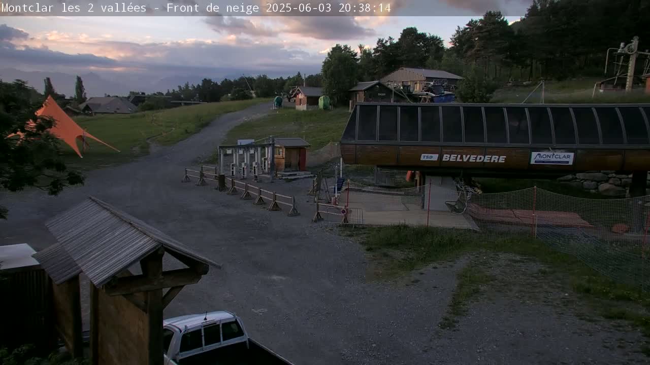 A gravel road leads to a chairlift station in a mountain setting at dusk, with a large orange tent and several small buildings nearby under a partly cloudy sky.