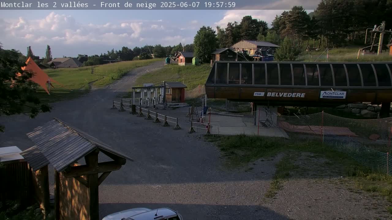 A chairlift station sits on a gravel path amidst green hills and a partly cloudy sky.