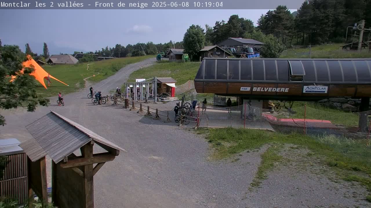 A mountaintop scene under a partly sunny sky shows a gravel path leading to a chairlift station with several mountain bikers near it, and a large orange fabric structure to the left.