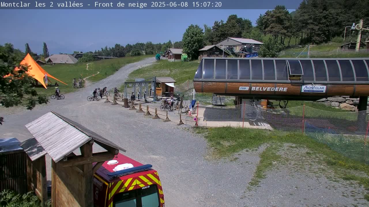 A mountaintop scene on a sunny day shows several mountain bikers near a chairlift, a large orange tent, and a firetruck.