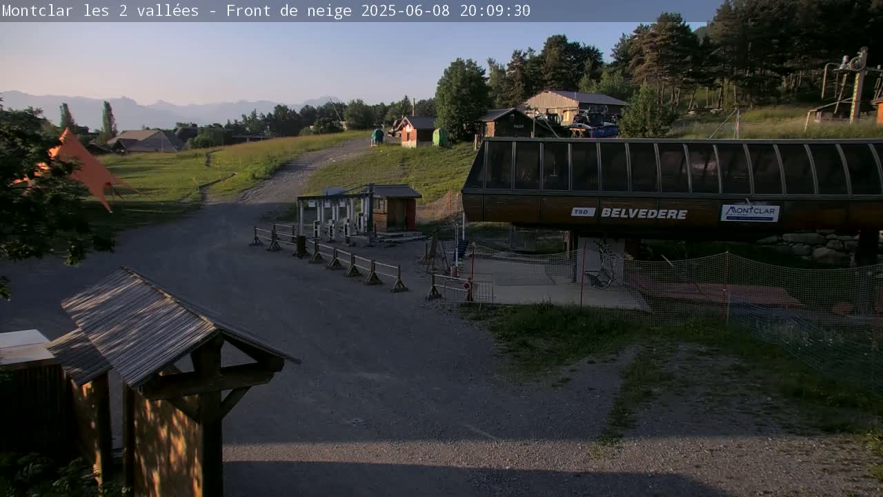 A chairlift station sits in a grassy area with a gravel path leading to it, under a clear evening sky and distant mountains.