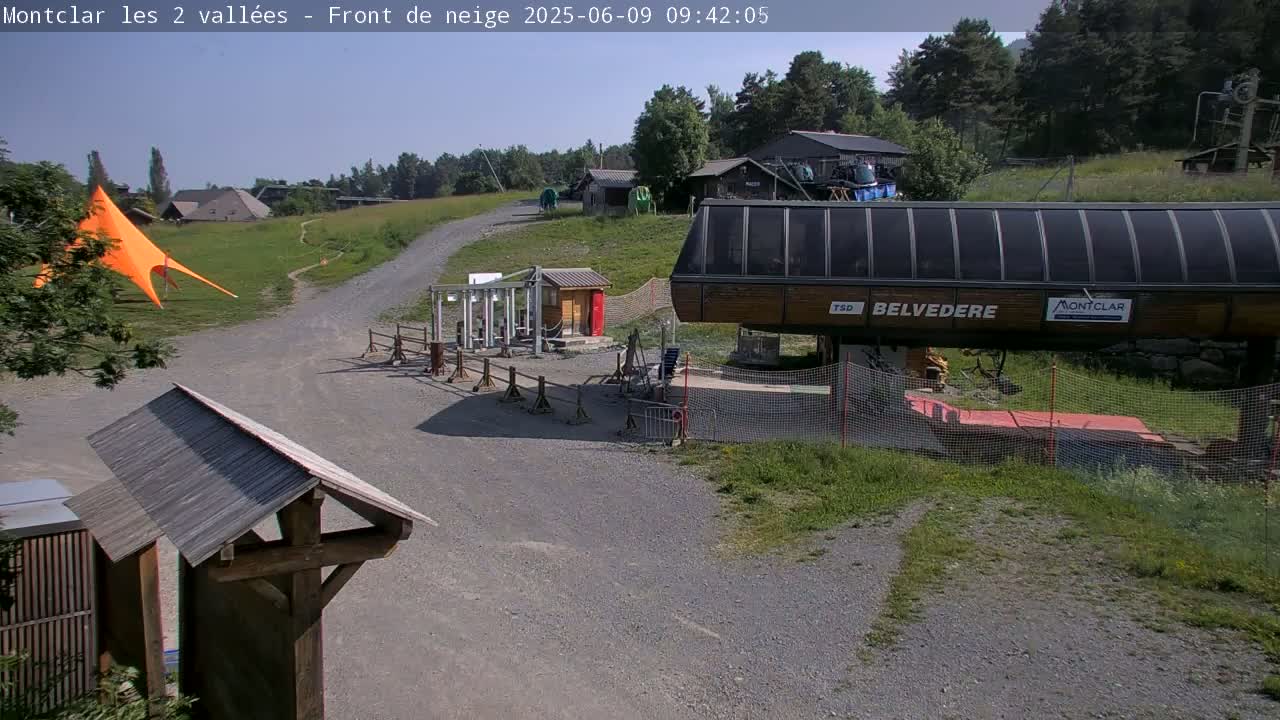 A chairlift station sits at the bottom of a grassy hill on a sunny day, with a large orange canopy and several small buildings nearby.